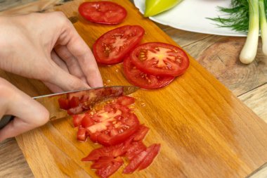 Kadın eli bıçakla kesme tahtasındaki domatesi bir tabak sebzenin yanında kesiyor.