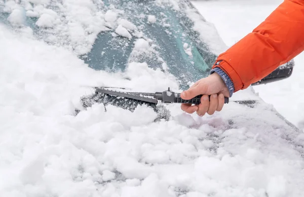 Man\'s hand in an orange jacket cleans snow from the car with a brush, a lot of snow on the car