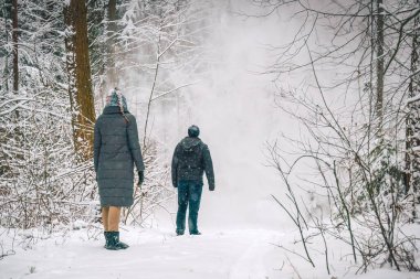 Genç bir kadın ve bir adam ormanda durup ağaçlardan düşen karı, arkadan çekilen fotoğrafı izliyorlar. Bir adam ve bir kızla güzel bir kar ormanı.