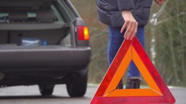 Bir kadın kaza yapmış bir arabanın yanında dururken acil durum levhası koyuyor..