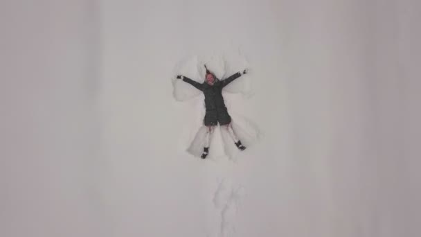 Uma mulher deita-se na neve e desenha um anjo. Tiroteio de um drone. — Vídeo de Stock