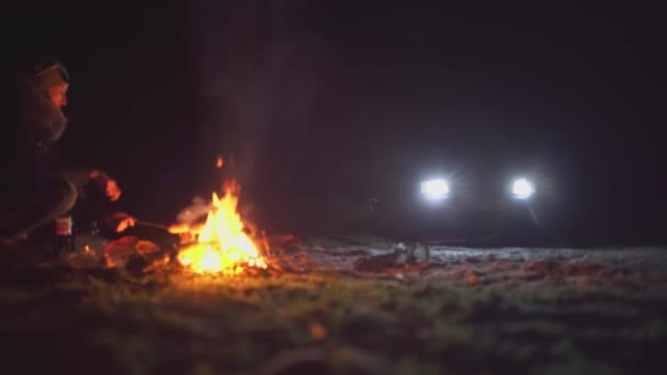 Kvinna vid lägerelden och stekt korv brinner på natten. bil med strålkastarna tända. — Stockvideo