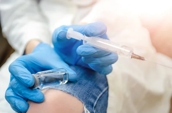 身穿白色实验室外套和蓝色手套的女医生手里拿着注射器 膝盖上的一瓶疫苗和一条破牛仔裤的特写 药品和疫苗接种的概念 — 图库照片