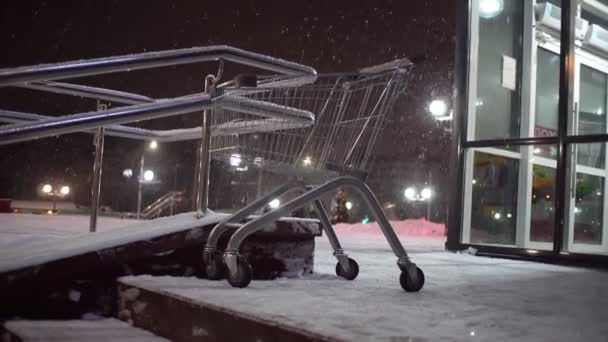 Een boodschappenkarretje staat 's nachts in een zware sneeuwval op de parkeerplaats van de supermarkt. — Stockvideo