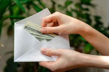 Kadının elinde içinde dolar olan beyaz bir zarf var. Arka planda bulanık bitkiler var. Kadın bir zarftan, maaştan, rüşvetten ya da tasarruftan para çekiyor.
