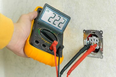 Erkek elektrikçi, beyaz duvardaki voltajı kontrol ediyor. Evin içinde multimetre var.