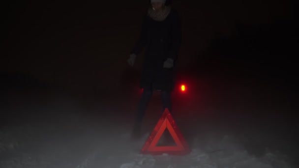 En flicka sätter upp en nödstoppsskylt bredvid sin bil på en vinternatt. — Stockvideo