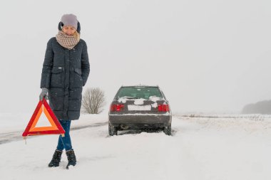 Ceketli, şapkalı, atkılı, ayakkabılı genç bir kız, bir arabanın arka planına karşı karla kaplı bir yolda uyarı işareti üçgeni koyuyor. Araba arızası kavramı, yardım bekliyor.