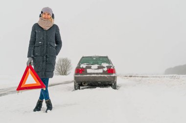 Ceketli, şapkalı, atkılı, ayakkabılı genç bir kız, bir arabanın arka planına karşı karla kaplı bir yolda uyarı işareti üçgeni koyuyor. Araba arızası kavramı, yardım bekliyor.