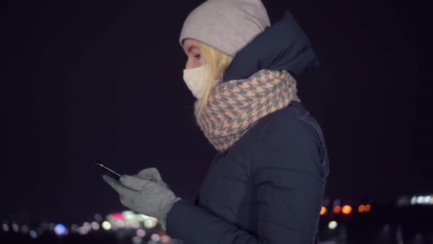Zamaskowana dziewczyna patrzy na telefon komórkowy w nocy na tle miasta. — Wideo stockowe