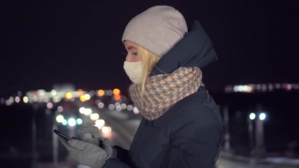 Μασκοφόρο κορίτσι κοιτάζει το κινητό της τη νύχτα στο φόντο της πόλης. — Αρχείο Βίντεο