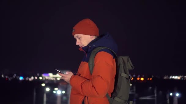 Um cara jovem olha para seu telefone celular à noite contra o fundo da cidade. — Vídeo de Stock
