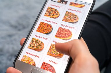 Kadın, ekranında pizzacı web sitesi olan cep telefonunu pofuduk gri bir battaniyenin üzerinde yatarken tutuyor. Kadın telefonla pizza sipariş ediyor.