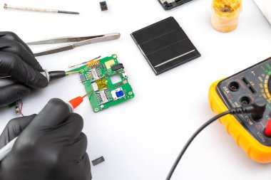 Radyo mühendisliği ustası cep telefonunun bir kısmını onarıyor, dijital çokölçer ile ölçümler yapıyor, siyah eldivenli ustanın elleri görülebiliyor.