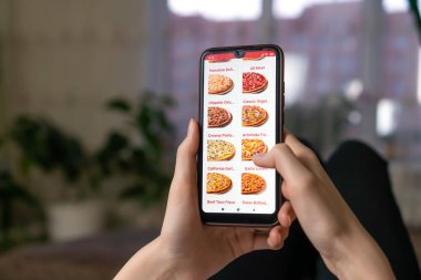 Kadın, ekranda pizzacının web sitesi olan cep telefonunu bir odada kanepede uzanmış halde tutuyor. Kadın telefonda pizza sipariş ediyor.