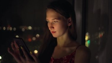 Genç ve güzel bir kadın gece pencere kenarında oturur ve telefonda mesaj yazar..