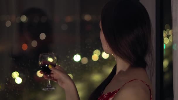 Woman stands at window with glass of wine in her hand watches night life of city — Stock Video