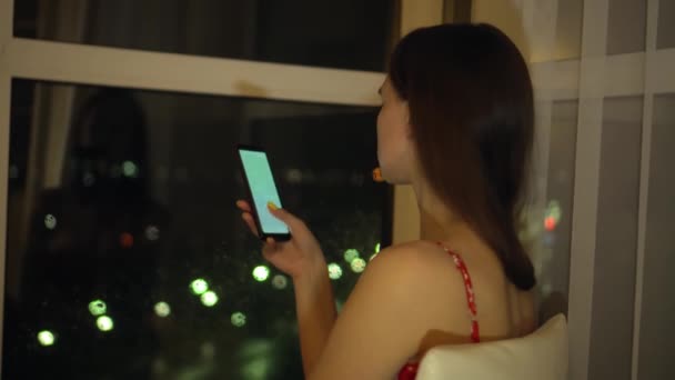 Menina segura o telefone e olha para ele na janela contra o fundo da cidade noturna. — Vídeo de Stock