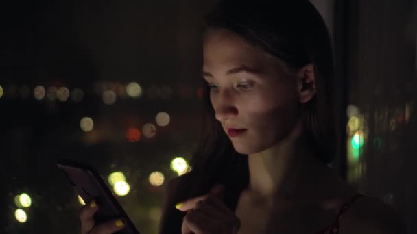 Ragazza tiene il telefono e lo guarda alla finestra sullo sfondo della città di notte. — Video Stock