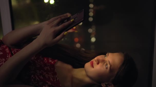 Girl holds phone and looks at it at window against background of night city. — Stock Video