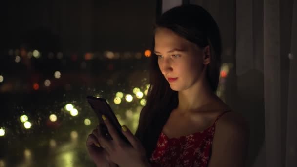 Девушка держит телефон и смотрит на него в окно на фоне ночного города. — стоковое видео