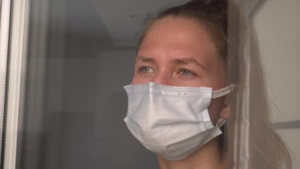 Egy fiatal nő orvosi védőmaszkban kinézett az ablakon. — Stock videók