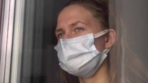 A young woman in a medical protective mask looks out the window — Stock Video