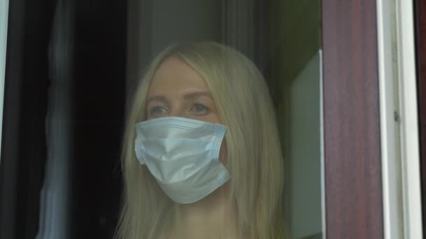 Uma jovem mulher com uma máscara protetora médica olha pela janela — Vídeo de Stock