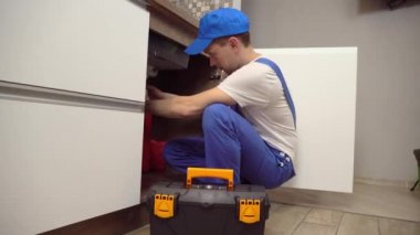 Tulumlu ve beyaz tişörtlü tesisatçı mutfaktaki boruları inceliyor..