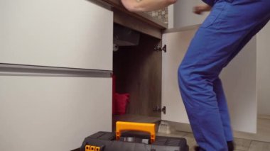 Tulumlu ve beyaz tişörtlü tesisatçı mutfaktaki boruları inceliyor..