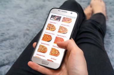 MOGILEV, BELARUS-23 Mart 2021: Kadın, pofuduk gri bir battaniyenin üzerinde yatarken ekranda bir pizzacının web sitesi ile yakın plan cep telefonu tutuyor. Kadın telefonla pizza sipariş ediyor.