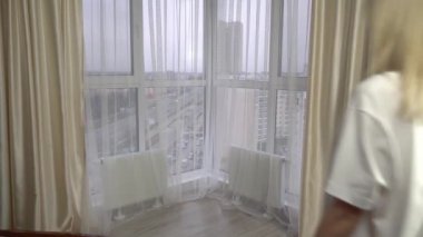 Kadın iç çamaşırlarıyla pencerenin önünde uzanan perdeleri açıyor.