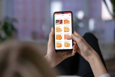 MOGILEV, BELARUS-23 Mart 2021: Kadın bir odada bir kanepede uzanırken ekranda pizzacının web sitesi olan bir cep telefonunun yakın çekimini tutuyor. Kadın telefonda pizza sipariş ediyor.