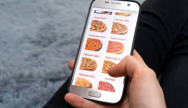 MOGILEV, BELARUS-23 Mart 2021: Kadın, ekranında pizzacı web sitesi olan bir cep telefonunu tutarken pofuduk gri bir battaniyenin üzerinde duruyor, fotokopi alanı. Kadın telefonla pizza sipariş ediyor.