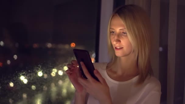 Genç beyaz kadın gece pencerenin önünde durmuş, telefonda haberleri okuyor.. — Stok video