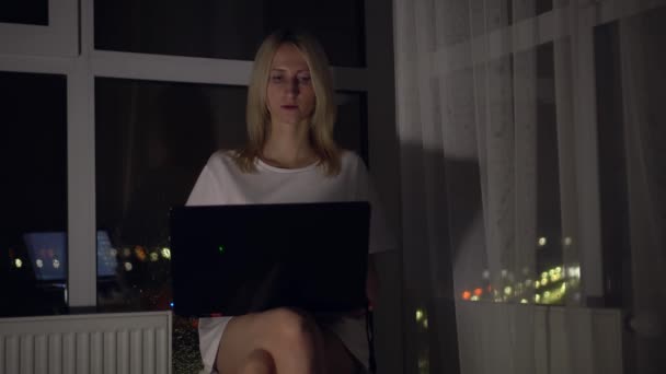 Женщина работает с ноутбуком, сидя у окна, на фоне ночного города. — стоковое видео