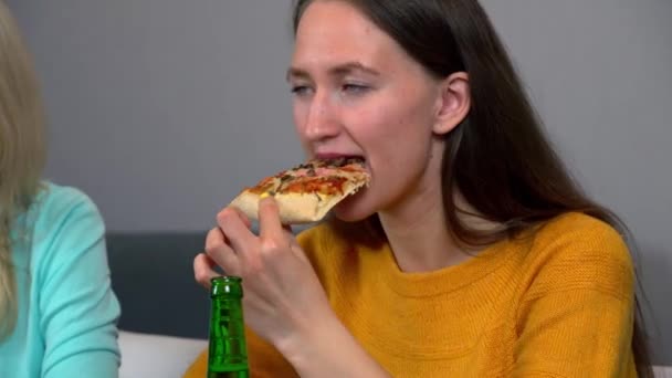 Két lány sört iszik és finom pizzát eszik otthon, a konyhájukban.. — Stock videók