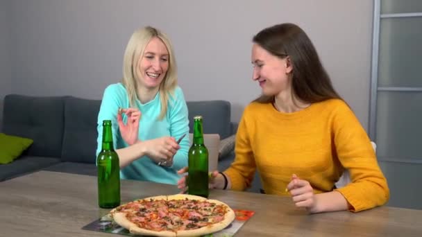 Две самые счастливые девушки дома на кухне едят пиццу и пьют пиво. — стоковое видео