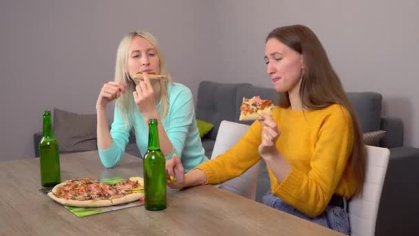 Дві дівчини, п'ють пиво і їдять смачну піцу, вдома на кухні . — стокове відео