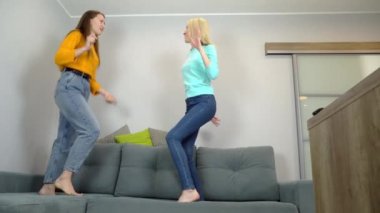 Açık renk ve siyah saçlı kadınlar oturma odasındaki büyük kanepede birlikte zıplıyorlar..