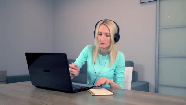 女性はヘッドフォンやヘッドセットを装着してノートパソコンで自宅で働いています。ノートを作る — ストック動画