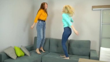 Açık renk ve siyah saçlı kadınlar oturma odasındaki büyük kanepede birlikte zıplıyorlar..