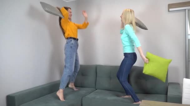 Två vänner eller systrar slåss med kuddar, hoppar på soffan hemma. — Stockvideo