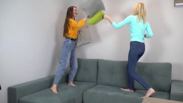 Dva přátelé nebo sestry se hádají s polštáři, skákání na gauči doma. — Stock video