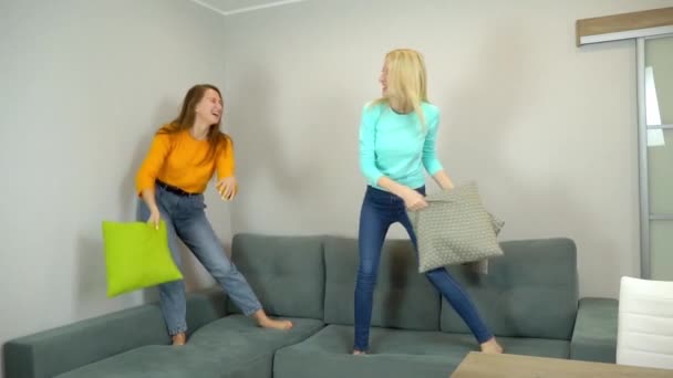 Two friends or sisters fight with pillows, jumping on the couch at home. — Stock Video