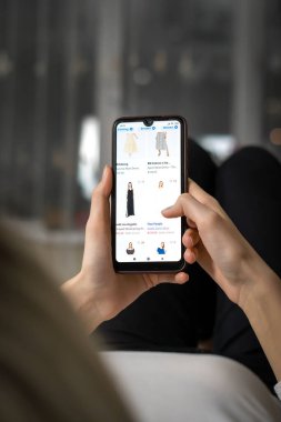 MOGILEV, BELARUS-23 Mart 2021: Kadın oturma odasındaki kanepede uzanıyor ve kıyafetlerini internet üzerinden yakın plan cep telefonu uygulaması üzerinden seçiyor. İnternetten alışveriş konsepti. Dikey fotoğraf