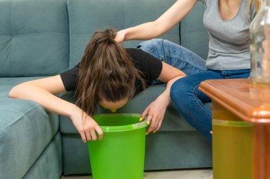 Koyu renk saçlı genç kadın kanepede yatıyor, alkol zehirlenmesinden hasta, bir kovanın üzerine kusacak, bir arkadaşı yardıma geldi, saçından tuttu, boş şişeler zehirlidir.