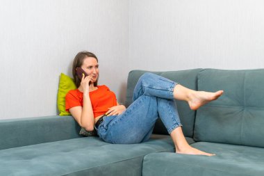 Koyu renk kotlu, turuncu gömlekli güzel bir kadın kanepede uzanıyor ve telefonda konuşuyor.