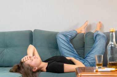 Sarhoş, genç, siyah saçlı bir kadın partiden sonra kanepede uzanıyor ve kafasını tutuyor, boş şişeler masanın üstünde.