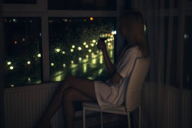 Beyaz gömlekli, elinde bir bardak kırmızı şarapla beyaz bir sandalyede oturan, tavandan zemin camına bakan, ince, sarışın genç bir kadın.